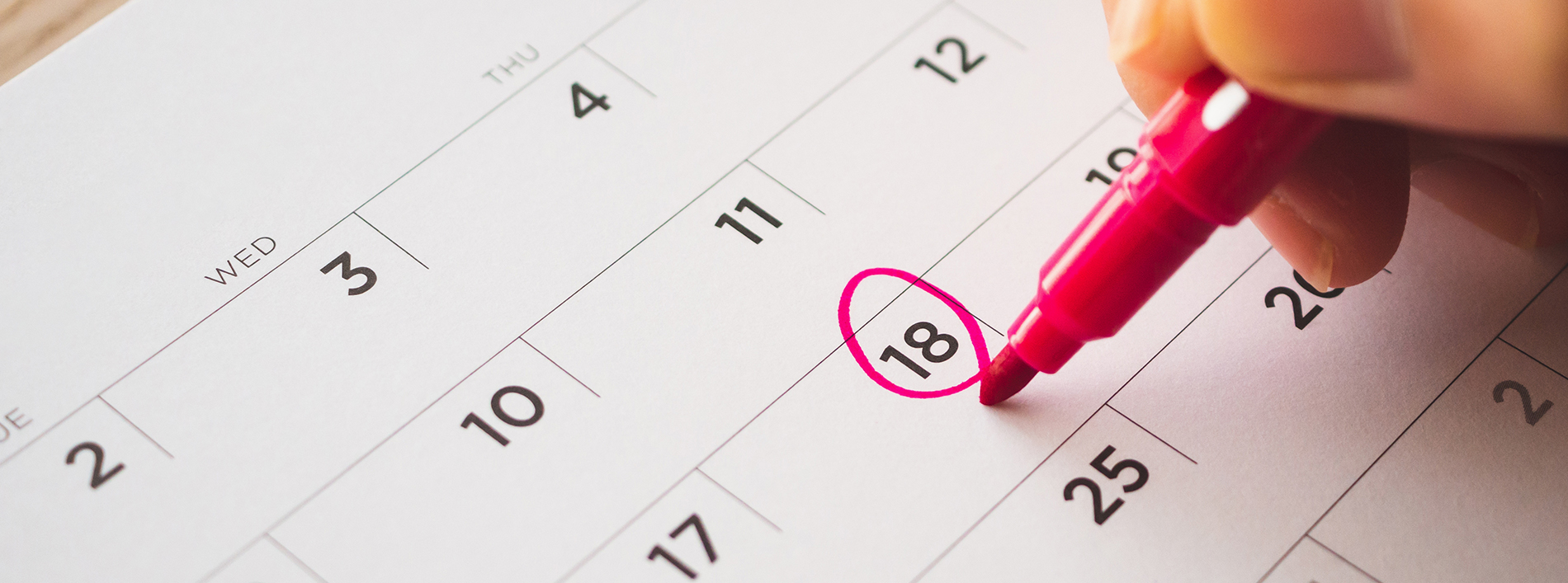 The image shows a close-up of a hand holding a pink highlighter marker, pointing to the number 18 on a printed calendar.