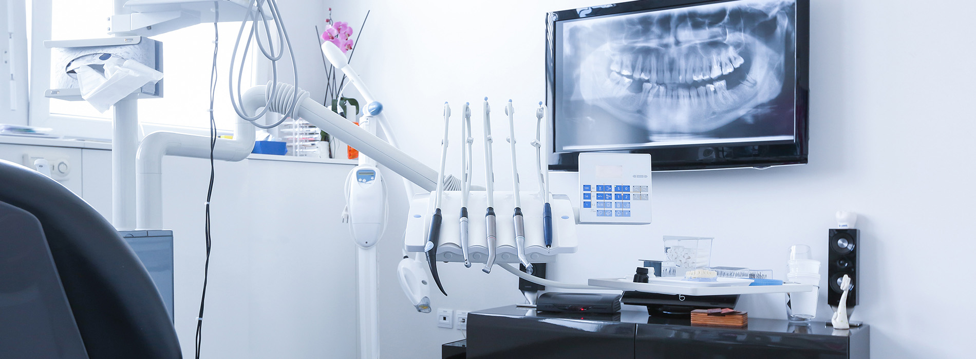 The image shows a modern dental office with a clean, white interior, featuring a dental chair and equipment, such as a monitor displaying an X-ray of a mouth, a dental light, and various other dental tools.