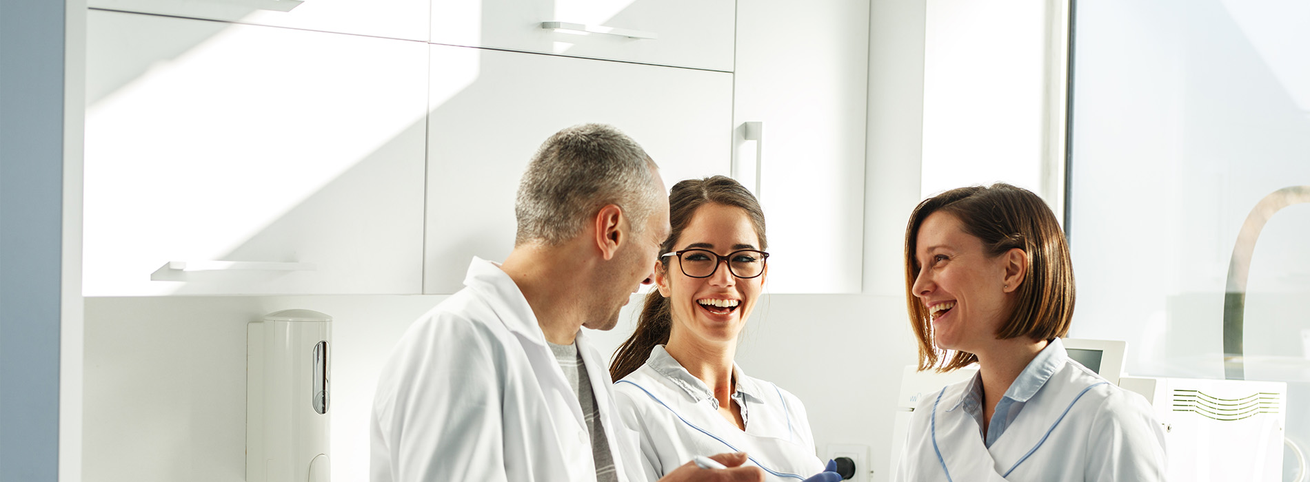 The image shows a group of four individuals in a professional setting, possibly a dental or medical office, with one person seated and three standing, engaging in conversation.