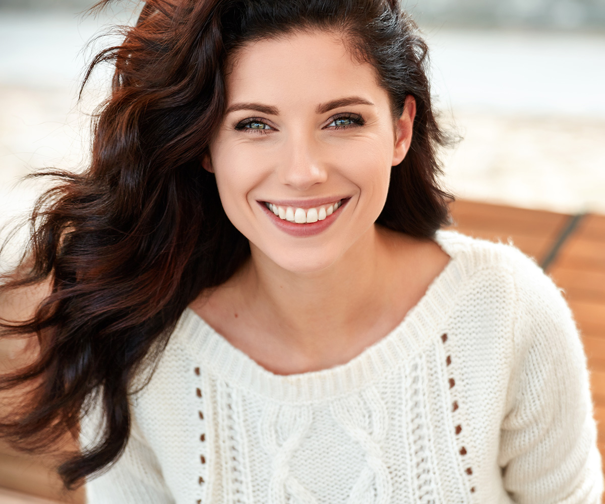 The image is a portrait of a woman with long brown hair, smiling at the camera. She has fair skin and appears to be in her late 20s or early 30s. The background is blurred but suggests an outdoor setting with natural light.