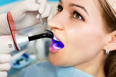 The image shows a person, likely a woman, sitting in a dental chair with her mouth open and a dental device inserted into her mouth. She is being attended to by a dental professional who is using an instrument to examine or treat the woman s teeth. The setting appears to be a dental office or clinic, indicated by the presence of medical equipment and the professional attire of the dental staff.