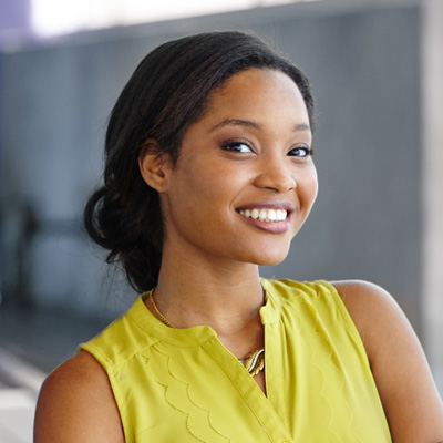 The image features a smiling woman with dark hair, wearing a yellow top and standing against a grey background. She has her left hand on her hip and is looking directly at the camera.