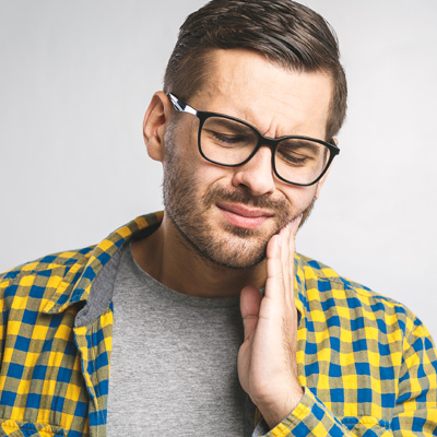 The image shows a young man with glasses, wearing a plaid shirt and holding his hand to his mouth. He appears to be in pain or discomfort.
