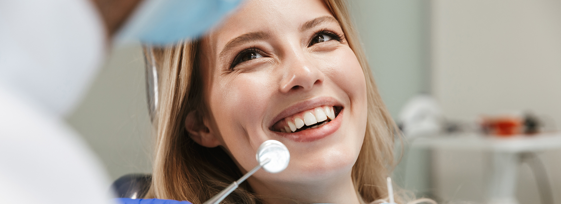 The image shows a woman smiling in front of a dental mirror, with a dentist wearing a mask and gloves behind her.