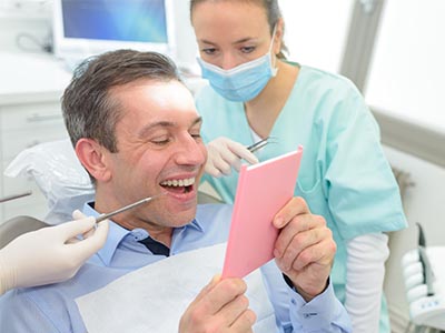A man sitting in a dental chair, holding a pink card, and smiling while a dentist is performing a procedure.