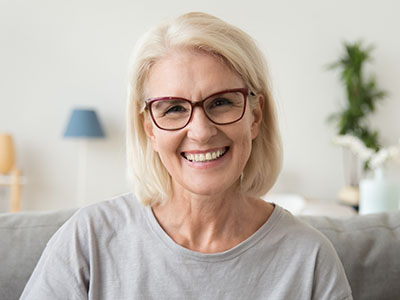 The image shows a woman with short blonde hair, wearing glasses and a light-colored top, smiling at the camera. She has white skin and appears to be indoors, possibly in a living room setting.