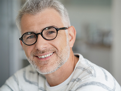 The image shows a smiling man with glasses, wearing a white shirt and looking directly at the camera.