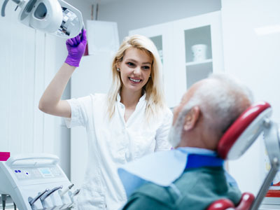The image shows a dental professional, likely a woman, standing next to an older man who is seated in a dental chair with his mouth open. She appears to be holding a device over his head and looking at him with a slight smile.