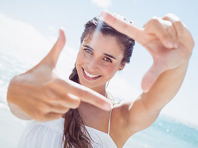 A woman with long hair is smiling while holding up a finger in front of her face, with the ocean behind her.