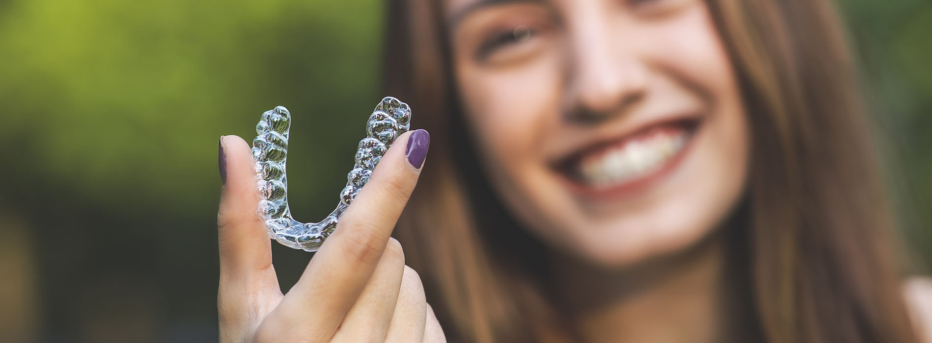 The image shows a person holding a clear plastic dental retainer, smiling at the camera.